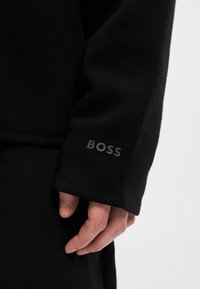 Μαύρο ενδύμα με μακρυά μανίκια και λείο υφή. Διαθέτει εμφανές λογότυπο "BOSS" σε σκούρο γκρι στη μανσέτα. Καθαρός και μινιμαλιστικός σχεδιασμός.