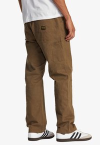 Khaki bomuldsbukser med en afslappet pasform, der har en baglomme og et logo-patch. Sammen med hvide sneakers med sorte striber.