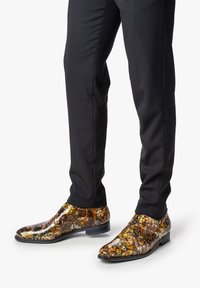 Chaussures de ville brillantes avec un motif texturé multicolore en brun, or et noir, associées à un pantalon noir slim.