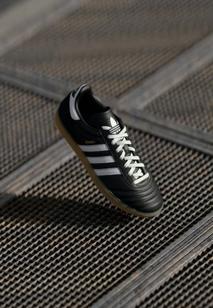 Czarne buty sportowe Adidas Samba z białymi paskami i sznurowadłami, brązową gumową podeszwą, spoczywające na teksturowanej metalowej powierzchni w formie kratki.