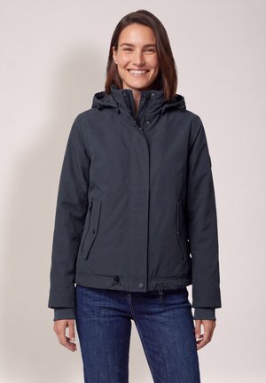Femme souriante portant une veste d'hiver sombre à capuche et un jean bleu, debout devant un fond clair uni.