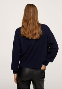 Donna con i lunghi capelli castani, che indossa un maglione blu navy e pantaloni in pelle neri, in piedi con una mano nella tasca posteriore, voltata di spalle.