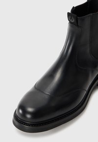 Bota de cuero negro hasta el tobillo con un acabado suave, puntera redondeada, paneles laterales elásticos y una pequeña etiqueta de logo en la parte trasera. Suela de goma.