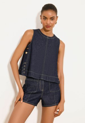 Vrouw die een mouwloos donker denimtopje draagt met knoopdetails aan de zijkant en bijpassende denimshorts met hoge taille, staande tegen een witte achtergrond.