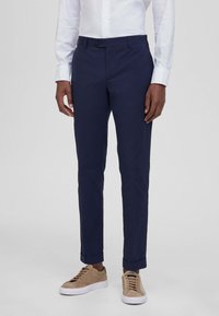 Pantaloni blu navy con orli risvoltati, realizzati in un tessuto morbido. Presentano una chiusura con bottone e tasche frontali. Indossati con sneaker beige.