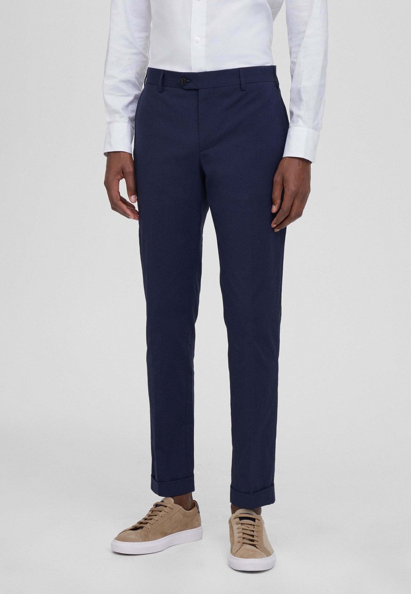 Pantaloni blu navy con orli risvoltati, realizzati in un tessuto morbido. Presentano una chiusura con bottone e tasche frontali. Indossati con sneaker beige.