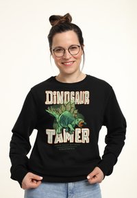 Felpa nera con grafica di un dinosauro colorato e testo "Dinosaur Tamer". Fit casual con polsini e orlo a coste.