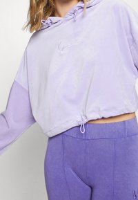 Hoodie corto lilla con cappuccio regolabile, caratterizzato da un logo ricamato a contrasto. Abbinato a leggings viola a vita alta, dalla texture liscia.