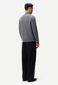 Mann trägt einen grauen Rollkragenpullover und lockere schwarze Hosen, steht mit dem Rücken zur Kamera vor einem hellen Hintergrund.