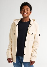 Chaqueta con capucha color crema hecha de tela suave, con botones de presión negros y bolsillos frontales, llevada sobre una camiseta negra y jeans azules.