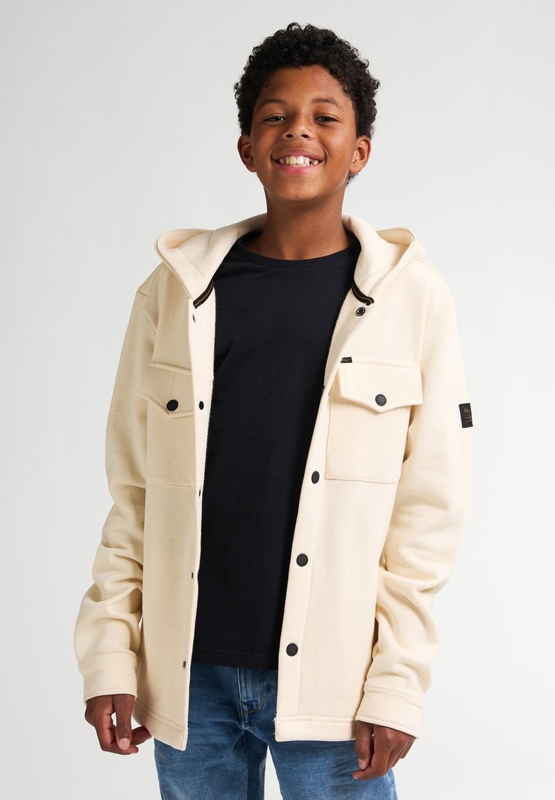 Chaqueta con capucha color crema hecha de tela suave, con botones de presión negros y bolsillos frontales, llevada sobre una camiseta negra y jeans azules.