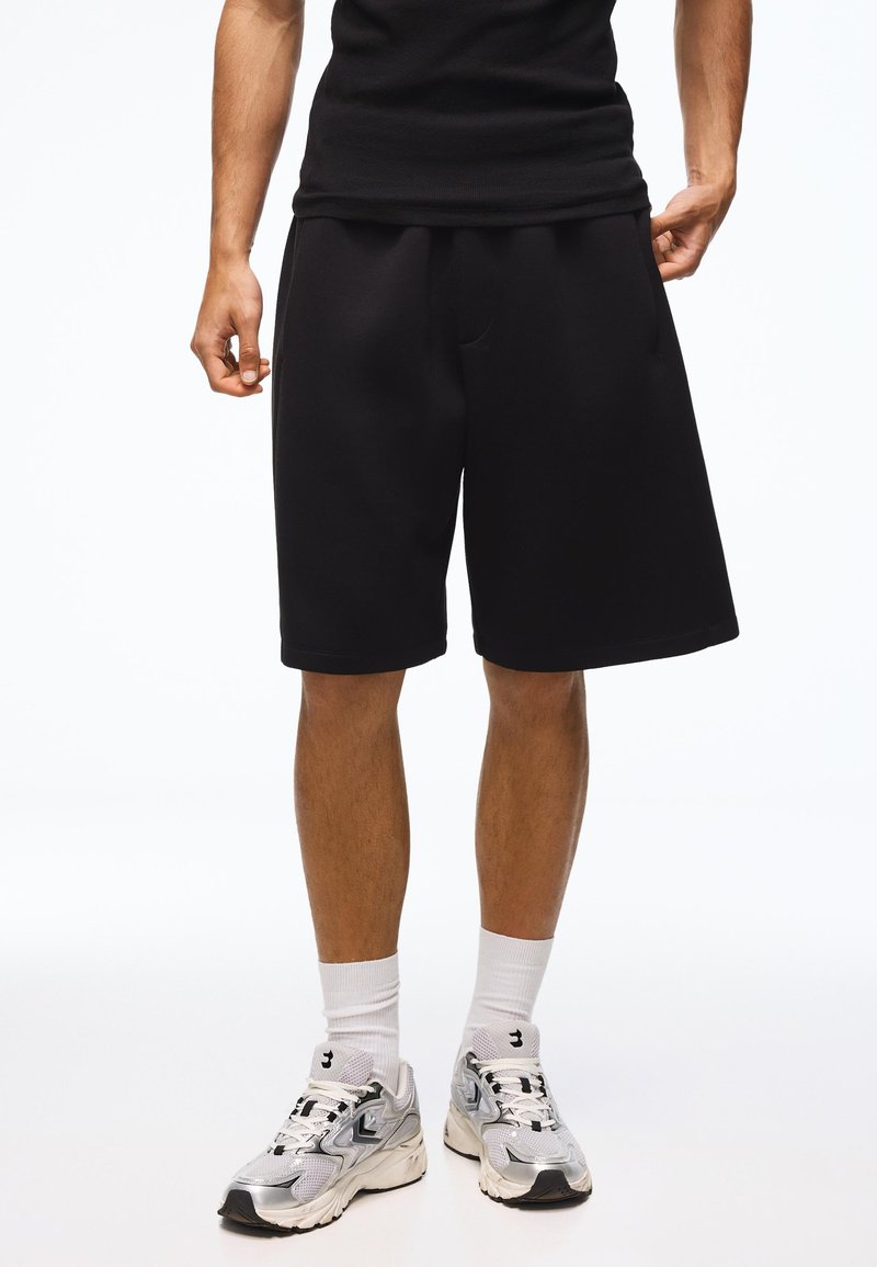 Homme portant un short noir mi-genou, des chaussettes blanches, des chaussures de sport argentées et blanches, et un t-shirt noir sur fond blanc.