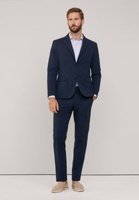 Marineblauer Anzug mit einem strukturierten Sakko und maßgeschneiderten Hosen, darunter ein gestreiftes Hemd. Beigefarbene Slipper runden das Outfit ab.