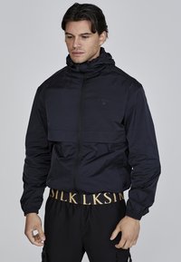 Jachetă windbreaker de culoare albastru marin, cu guler înalt, fermoar frontal și manșete elastice. Prezintă un buzunar orizontal la piept și un design elegant.