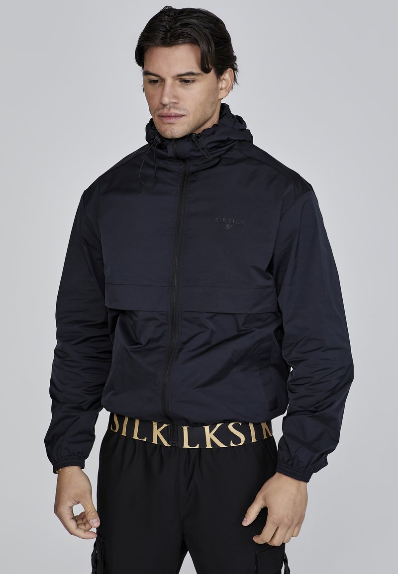 Jachetă windbreaker de culoare albastru marin, cu guler înalt, fermoar frontal și manșete elastice. Prezintă un buzunar orizontal la piept și un design elegant.