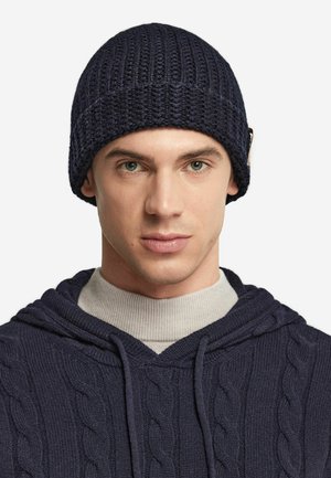 Marineblaue Strickmütze mit geripptem Muster. Begleitet von einem grauen Rollkragenpullover und einem marineblauen Kapuzenpullover mit Zopfstrick, der eine Kapuze und einen Kordelzug hat.