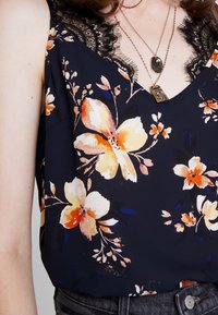 Blommig blus i marinblått tyg med orange och gula blommor. Har spetsdekoration vid halslinningen. Lätt material, avslappnad passform.