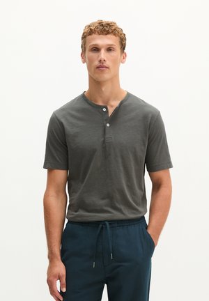 Jonge man met krullend haar, dragend een donkergrijze Henley met korte mouwen en donkerblauwe broek met trekkoord, staand met één hand in de zak.