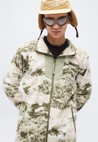 Νεαρός που φοράει ανακλαστικά γυαλιά ηλίου, καφέ χειμερινό σκουφί με καλύμματα αυτιών και πράσινο-μπεζ ζακέτα από φλις με φερμουάρ και σχέδια.