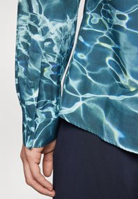 Camicia button-up in teal con un motivo acquatico. Maniche lunghe, un polsino con bottone e orlo dritto. Tessuto liscio con una leggera lucentezza.