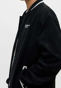 Black bomber jacket made of soft fabric, featuring white striped ribbed collar and cuffs, with embroidered text on the chest and subtle patterns.