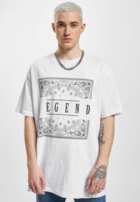 Hvit bomulls t-skjorte med svart paisley-mønster rundt og ordet "LEGEND" i fete bokstaver, brukt sammen med dongeri-bukser.