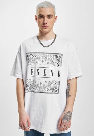 Weißes Baumwoll-T-Shirt mit einem schwarzen Paisleymuster-Rahmen und dem Wort "LEGEND" in fetten Buchstaben, getragen mit Jeans.