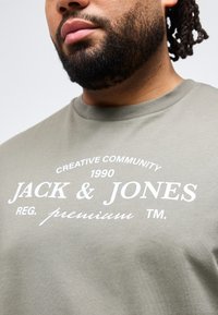 Άνδρας με γενειάδα που φοράει ελιάς πράσινη μπλούζα με λευκό κείμενο "JACK & JONES premium" και τύπωμα "Creative Community 1990".