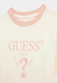 Krátký rukáv krémového trička s světle růžovým žebrovaným límcem. Obsahuje vyšívané logo "GUESS U.S.A." a dekorativní růžové akcenty.