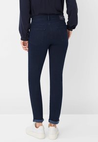 Donkerblauwe, slim-fit jeans met opgerolde boorden, achterzakken en een gladde textuur, gecombineerd met witte sneakers voor contrast.