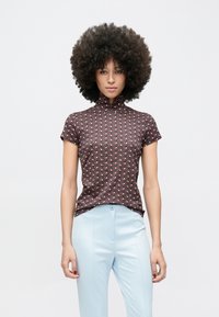 Top s krátkym rukávom z tmavej tkaniny s geometrickým vzorom v červenej a bielej farbe. Dizajn s vysokým golierom a priliehavým strihom. Kombinovaný so svetlomodrými nohavicami.