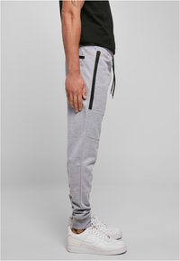 Pantalon de jogging gris avec poches zippées noires, taille à cordon de serrage et ourlets côtelés, porté avec des baskets blanches. Texture de tissu mat.
