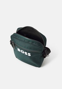 Grøn stof crossbody taske med lynlås på toppen, der har en frontlomme og hvidt logo. Inkluderer en justerbar rem og struktureret overflade.