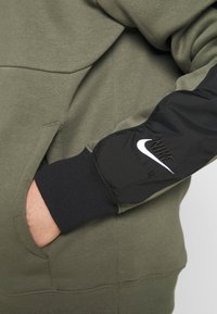 Sudadera verde oliva con una manga negra, que presenta un logo blanco de Nike y el texto "AIR". Textura suave, con un bolsillo tipo canguro visible.