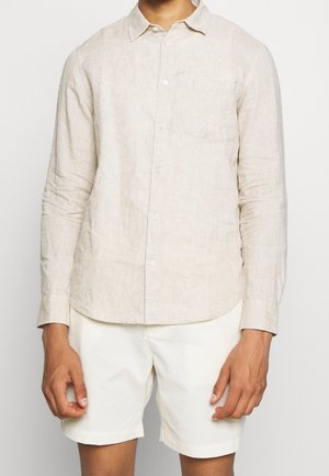 Man wearing long-sleeve beige linen button-up shirt and white shorts standing against a plain background.