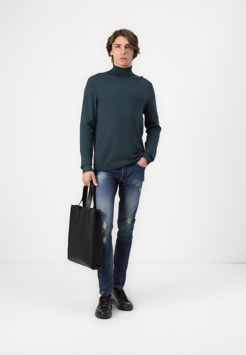 Un pull col roulé long à col monté en bleu sarcelle foncé associé à un jean bleu déchiré et un sac fourre-tout noir. Des chaussures noires à lacets complètent le look.