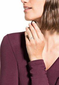 Anello d'argento con design intrecciato, dotato di piccole pietre trasparenti. Il modello ha capelli di lunghezza media e indossa un top a maniche lunghe color bordeaux.