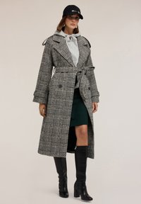 Trench coat scozzese in bianco e nero, con cintura in vita e grandi rever. Abbinato a un felpa grigia e una gonna verde, completato da stivali neri al ginocchio.