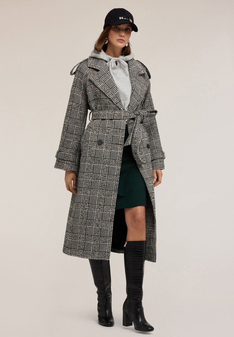 Trench coat scozzese in bianco e nero, con cintura in vita e grandi rever. Abbinato a un felpa grigia e una gonna verde, completato da stivali neri al ginocchio.