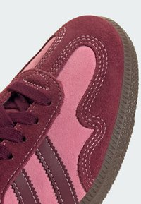 Sneaker mit bordeauxroten und rosa Wildlederpaneelen, kontrastierenden Nähten, texturierter Gummisohle und klassischem Drei-Streifen-Design.