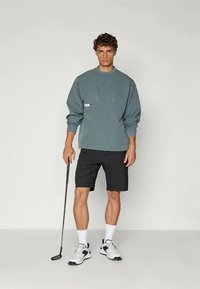 Hele roheline sweatshirt kahe tõmblukuga taskuga, koos mustade lühikestega. Mudel hoiab golfikeppi, kandes valgeid sokke ja spordijalatsit.