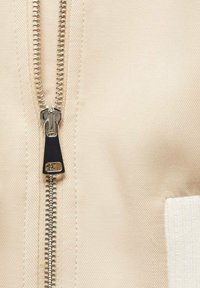 Bežová tkanina z blízka se stříbrným zipem s černým úchytem, vedle bílé ribbené tkaniny jako akcent. Hladký povrch a čisté linie.