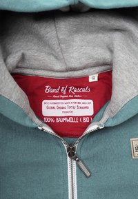 Huvtröja med dragkedja i teal och grått bomull. Har en grå inre foder, framdragkedja och en etikett som anger 100% ekologisk bomull.