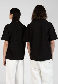 Tricouri negre din bumbac cu mâneci scurte, croi lejer și guler rotund; purtate cu pantaloni albi cu croi larg; branding minim vizibil pe laterale.