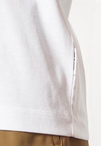 White cotton t-shirt with a side seam tag, displayed at an angle to show the texture and structure. Stitching is visible along the hemline.