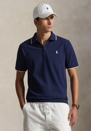 Muškarac u mornarskoplavoj polo majici s bijelim obrubom i logom, bijelim kratkim hlačama s vezicom, bijelom kapom s logom i smeđim ručnim satom, stoji s rukama u džepovima.