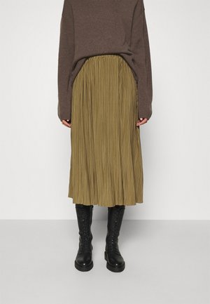Personne portant un pull marron, une jupe midi plissée vert olive et des bottes de combat noires à lacets, debout devant un fond uni clair.