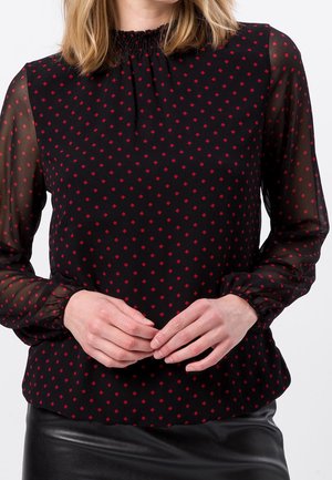Femme portant un chemisier noir transparent à manches longues avec un motif losange rouge et une jupe en cuir noire, mains jointes devant.