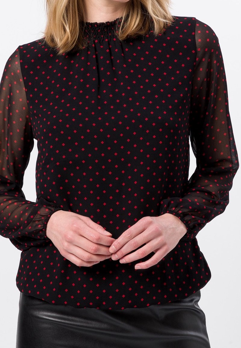 Femme portant un chemisier noir transparent à manches longues avec un motif losange rouge et une jupe en cuir noire, mains jointes devant.