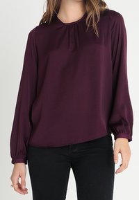 Femme portant un chemisier à manches longues, de couleur violet foncé, avec un col et des poignets froncés, associé à un pantalon noir ajusté.